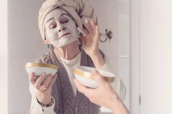 Kafasında havlu olan yaşlı bir kadın gülümseyip maske takıyor. .