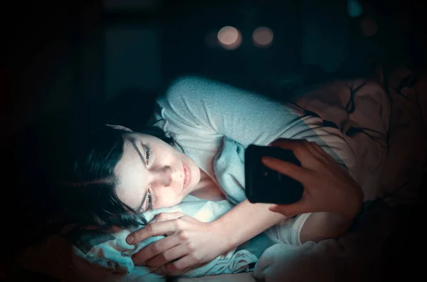 Молода дівчина в ліжку з телефоном вночі . Стокова Картинка