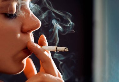 Genç bir güzel kız sigara içiyor, bir kadın stresli ve elinde bir sigara tutuyor, evinin penceresinin yanında duruyor, yakın çekim manzarası var..
