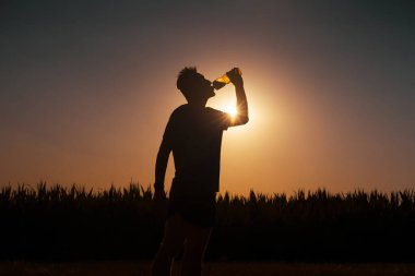 Genç adam egzersiz yaparken ve koşarken ferahlatıcı su içer. Güneşin arka planında bir şişe suyu olan bir sporcunun silueti..