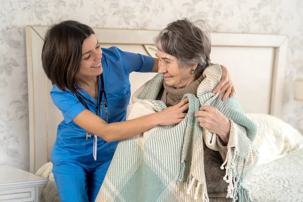 A young nurse takes care of an elderly 80-year-old woman at home, wraps a blanket around her. Happy retired woman and trust between doctor and patient. Medicine and healthcare.