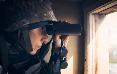 Askeri üniformalı ve kask takan genç adam savaş sırasında dürbünle bakıyor ve askeri tatbikatlara katılıyor. Asker binanın içerisindeki pencerenin yanında keşif yapıyor..