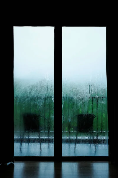 雨に濡れるガラス窓の暗いフレーム雨の季節のリゾートベッドルームの外の芝生と緑の木々の椅子のぼやけた背景 — ストック写真