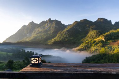 Relax corner of with lamp on wooden table with blurry background of mountain range at sunrise. White fog cover the hill and sunlight shining to mountain slope.