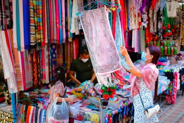 Chiangmai, Tayland - 1 Ocak 2022: Maskeli kadın Tayland 'ın kuzey kesiminin geleneksel ürünlerinin satıldığı caddedeki yerel bir dükkanda el yapımı renkli eşarp seçiyor.