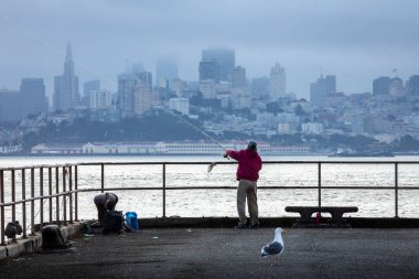 San Francisco, Kaliforniya, ABD - 27 Eylül 2019: Kırmızı ceketli son sınıf öğrencisi San Francisco 'nun puslu şehriyle denize balık oltası fırlattı.