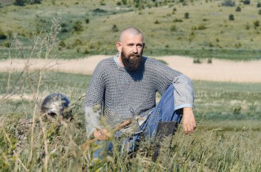 Viking. Keten gömlek üzerine metal zincirli kel sakallı adam elinde baltayla tepede oturuyor. Yakınlarda bir kask var. Kameraya bakıyorum..