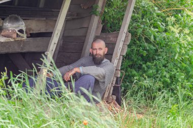 Viking savaşçısı savaştan sonra tahta binanın yakınında dinleniyor. Metal zincir zırh ve kask. Yeşil çalılar.