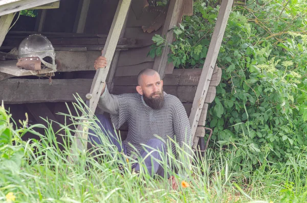 Viking savaşçısı savaştan sonra tahta binanın yakınında dinleniyor. Metal zincir zırh ve kask. Yeşil çalılar.