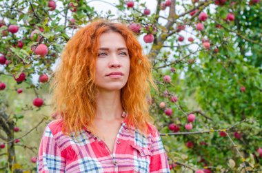 Elma bahçesinin bulanık arka planında güzel bir kadının parlak fotoğrafı. Kızıl saçlı, mavi gözlü, dalgın bakışlı.