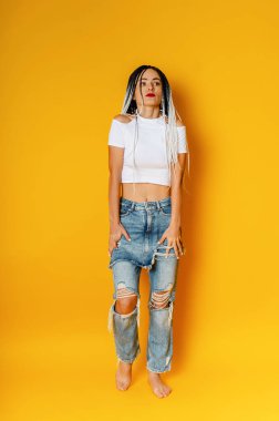 Saçları örgülü genç bir kadının fotoğrafı. Dreadlock 'lar. Beyaz tişört ve yırtık kot pantolon. Sarı stüdyo arka planı.