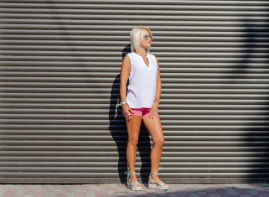 Güneş gözlüklü zayıf sarışın. Şort ve tişört giymişti. İnce, çıplak, uzun bacaklar. Güzel bir kadının sıcak yaz fotoğrafı. Yan görünüm