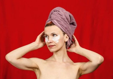Kafasında havlu olan yarı çıplak bir kadın. Ellerini başının üstüne koy. Gözlerinin altında kozmetik yamalar. Başka tarafa bakıyor. Göz çevresinde cilt bakımı. Kozmetoloji. Kırmızı arkaplan.