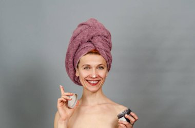 Kafasına havlu sarılmış, gri arka planda soyutlanmış neşeli, yarı çıplak kadın portresi. Gülümsüyor, krema sürüyor..