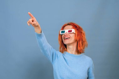 Cheerful young woman in 3D glasses. She points her finger up. Blue background