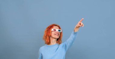Cheerful young woman in 3D glasses. She points her finger up. Horizontal photo. Blue background