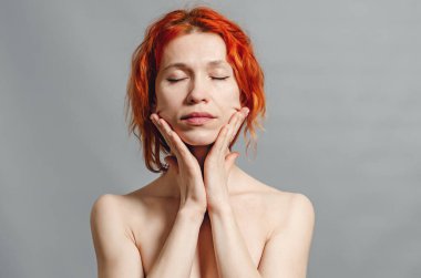 Kızıl saçlı çıplak kadın. Yüze yakın eller. Gözler kapalı. Rahat ol. Gri arkaplan.