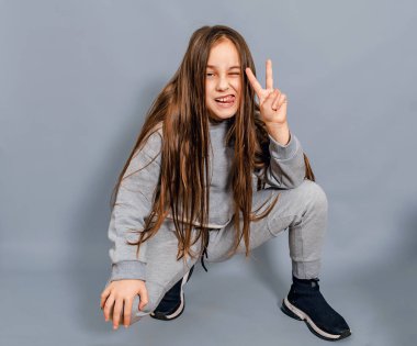 Uzun saçlı ve parmaklarıyla zafer işareti gösteren bir kız. Dilini gösteriyor, bir gözünü kıstı. Gri arkaplan.