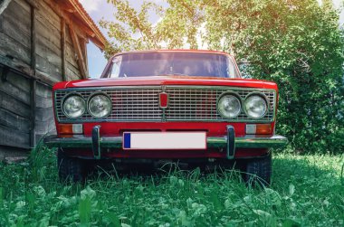 Front view of red vintage Lada car in countryside. Old wooden barn from side. Old red Soviet car.