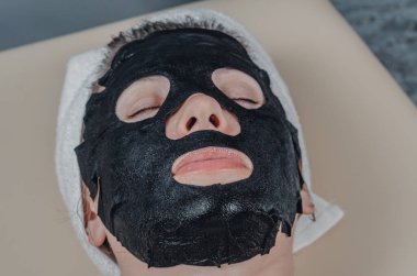 Black cosmetic fabric mask on woman's face. Face peeling mask with coal, spa procedures, skin care, cosmetology. Closed eyes. View from below