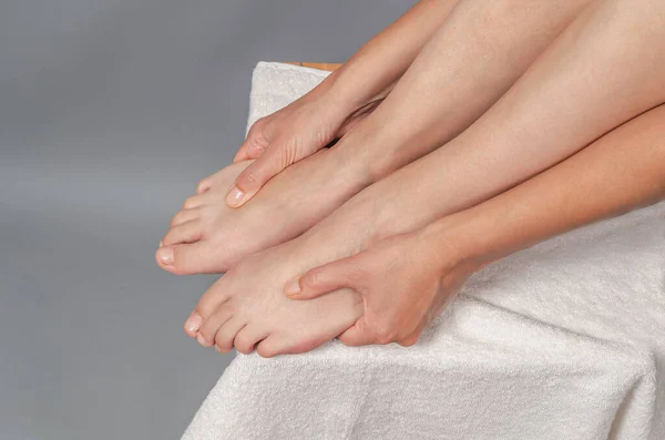 Point self-massage of feet. Woman touches her smooth legs, close-up. Legs and hands on white towel. Gray background.