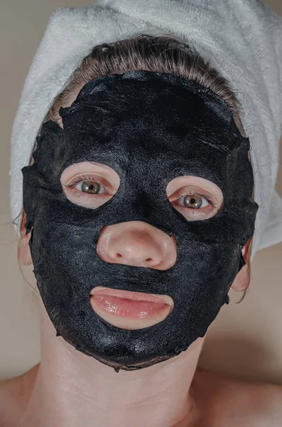 Black cosmetic fabric mask on woman\'s face. Face peeling mask with coal, spa procedures, skin care, cosmetology. Looks into camera.