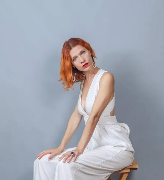 穿着白色衣服的红头发女人坐在木椅上 看着摄像头 红色口红 很深的领口手放在膝盖上 — 图库照片