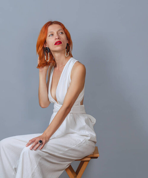 Red-haired woman with dreamy expression, focused to side, wearing elegant white dress, has red hair. Posing on gray background.