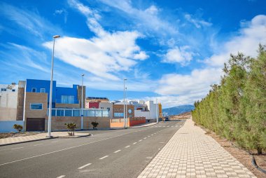 Deniz kıyısında kaldırımlı yol manzarası var. Mountainside ve iskeleye yakın binalar mavi gökyüzüne karşı arkasında beyaz bulutlar.