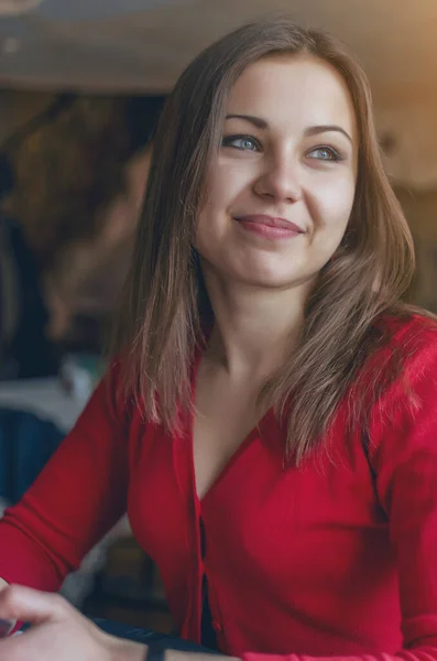 Cute brunette girl in red blouse is sitting at table in a cafe