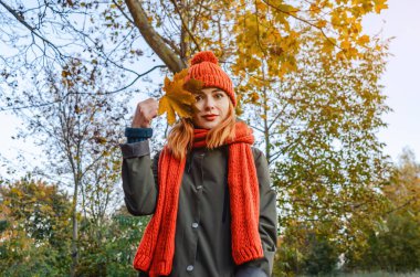 Elinde turuncu örgü şapkalı ve atkılı, güz sarısı yapraklı gizemli kadın. Sonbahar ilhamı.