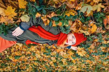 Sonbahar sarısı yaprakları elinde turuncu örgü şapka ve parktaki yaprakların üzerinde duran eşarpla rüya gibi bir kadının üst görüntüsü.