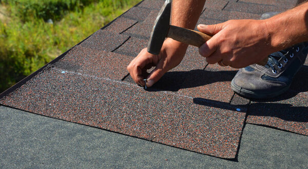 A close-up of asphalt shingles installation on the roof edge. A roofer is nailing asphalt shingles to the roof deck covered with roofing underlayment. Roofing construction.  