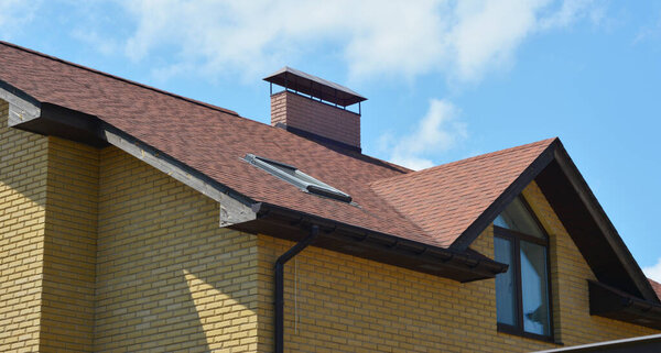 A close-up of a brick house with a brown asphalt shingled roof, soffit and fascia, skylight, chimney, attic skylight window and roof gutter system with a downspout. 