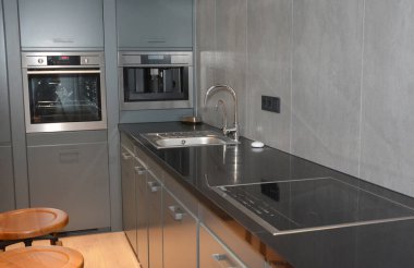 Modern bir mutfak iç tasarımı. Lavabosu ve elektrikli ocağı olan siyah bir mutfak tezgahı var. Gri dolaplar, dahili fırın, kahve makinesi ve ahşap döşemesi var..