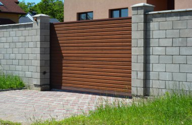 A close-up of a wood slat automatic sliding gate, entrance driveway sliding gate, gray brick fence with pillars, and paver driveway in front of a house.   