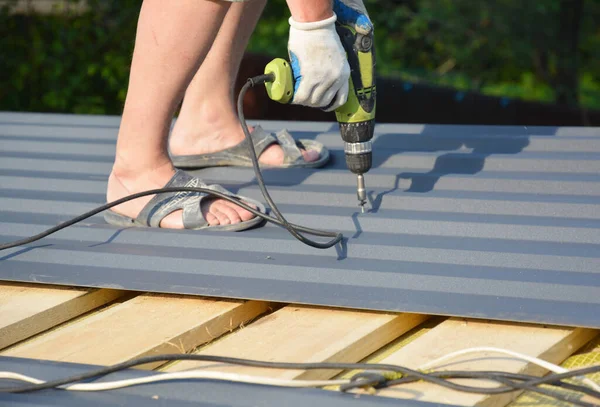 Metal roofing construction. A building contractor is installing corrugated metal roofing sheets on the roof rafters,  screwing down metal panels using electric screwdriver. 