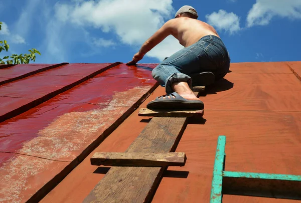 Painting old metal roof with a paint brush. Worker repainting house steel roof with red paint.