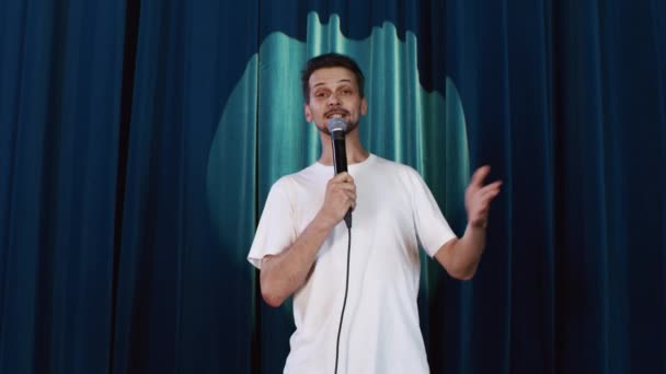 Comic man prezentuje stand-up na scenie z mikrofonem przeciwko kurtynie — Wideo stockowe