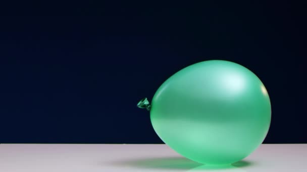 Red dart blows up green balloon lying on white table against blue background — Stock Video