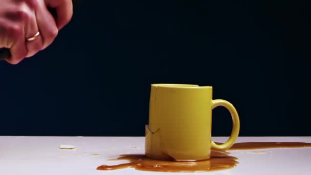 Рука чоловіка розбиває жовтий чайний кухоль рідиною з залізним молотком — стокове відео