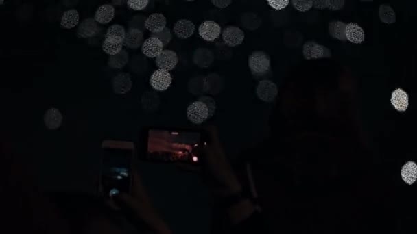 As mãos seguram smartphones e filmam vídeos de fogos de artifício explosivos no céu noturno. — Vídeo de Stock