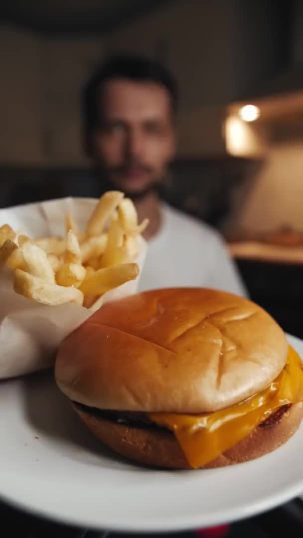 A man eats french fries next to a fast food cheeseburger on a white plate — Video Stock