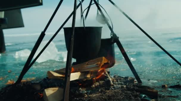 On fire with firewood food is cooked in large metal vats on frozen bank of river — Stock video
