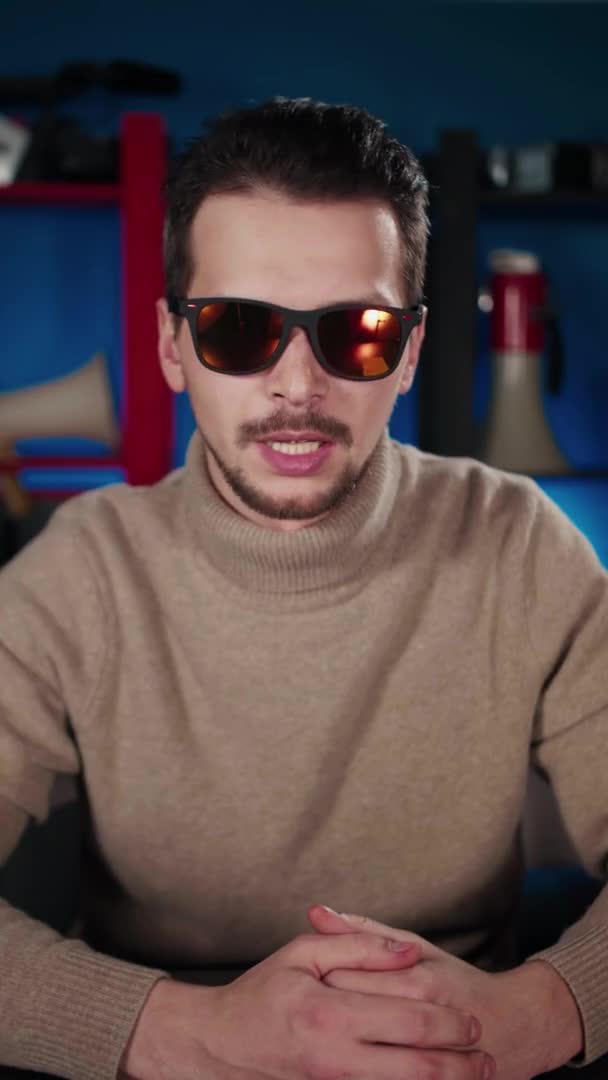 Ein Europäer mit Sonnenbrille sitzt an einem Tisch im Studio und sagt etwas — Stockvideo