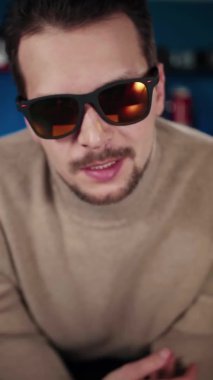 Güneş gözlüklü bir Avrupalı stüdyoda bir masada oturuyor ve bir şeyler söylüyor.