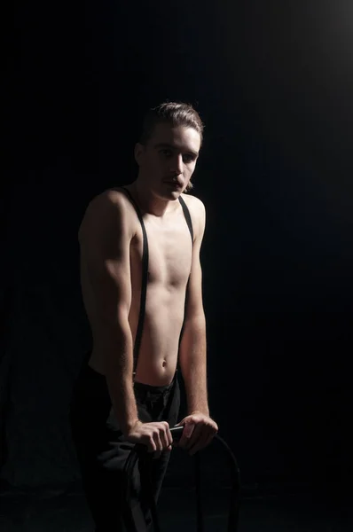 Çekici genç bir sanatçı, bıyıklı, pantolon askısı takmış, kameraya bakıyor ve tişörtsüz, klasik görünümlü bir sandalyeye yaslanıyor. Kabare ve LGBTQA kavramı