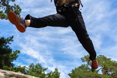 Dağınık bulutlar ve ağaçlarla dolu mavi gökyüzü ile kayaların arasında zıplayan bir yürüyüşçünün bacak görüntüsü..