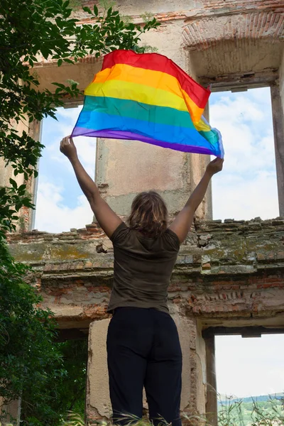 Sırtı dönmüş, kollarını kaldıran ve rüzgarda sallanan gökkuşağı bayrağını tutan yetişkin sarışın kadın. Pencerelerden gökyüzünü görebileceğin harabe bir binada. LGBTIQ Konsepti