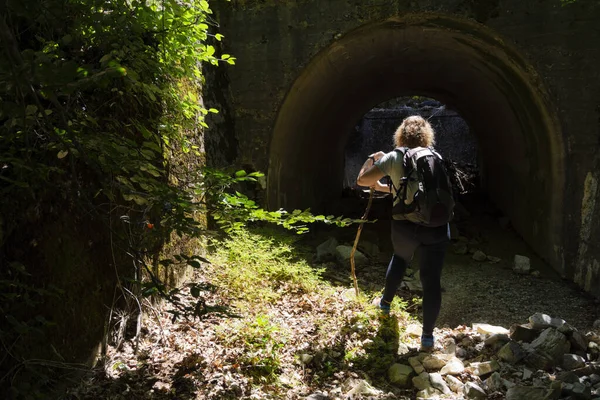 Sırtında sırt çantasıyla sarışın bir kadın yürüyüşçü, tahta bir direğe yaslanmış, kayın ormanının karşısındaki bir köprünün altından karanlığa bakıyor..